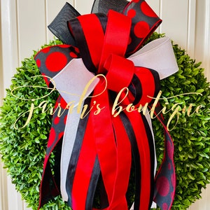 Puede incluir: Una corona de boj verde con un gran lazo de lunares rojos, negros y blancos. El lazo tiene múltiples capas de cinta y está atado con una cinta dorada.