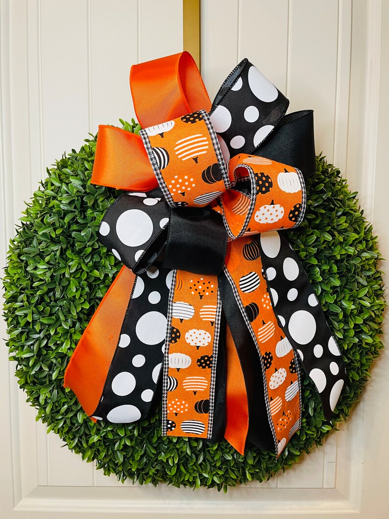 K&ouml;nnte beinhalten: Ein gr&uuml;ner Buchsbaumkranz mit einer gro&szlig;en Schleife aus schwarzem und orangefarbenem Band. Das Band hat ein wei&szlig;es Polka-Dot-Muster und ein Muster aus wei&szlig;en K&uuml;rbissen auf orangefarbenem Hintergrund.