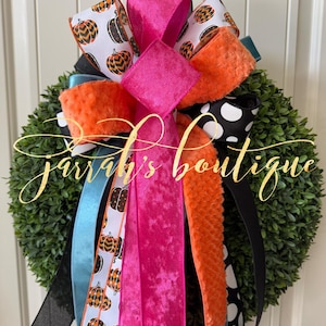 May include: A decorative wreath with a green base, adorned with colorful ribbons. The ribbons include patterns of pumpkins, polka dots, and solid colors like pink, orange, black, and teal. The text "Jarrar's boutique" is visible.