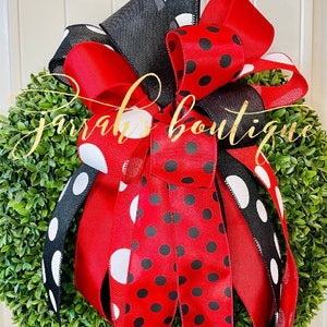 May include: A round green boxwood wreath adorned with a large decorative bow featuring red and black ribbons. The ribbons display various patterns including black polka dots on red, white polka dots on black, and solid red and black textures.