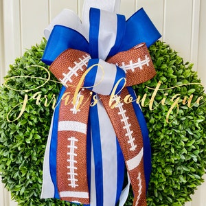 May include: A green boxwood wreath with a blue, white and brown football-themed ribbon bow. The ribbon has a white stripe and a brown football pattern.