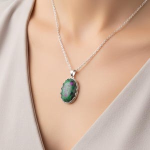 Puede incluir: Collar de plata con colgante ovalado. El colgante tiene una piedra verde y roja, enmarcada por pequeñas piedras transparentes. El collar lo lleva una persona con una camiseta gris claro. La cadena tiene un diseño delicado.