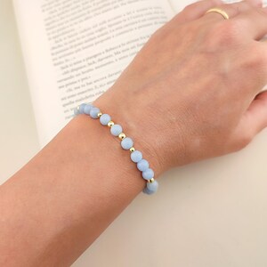 Peut inclure: Un bracelet de perles bleu clair avec des accents dorés est porté au poignet. Le bracelet est composé de petites perles rondes bleu clair entrecoupées de perles dorées. Une bague dorée est sur le doigt.
