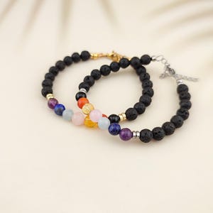 Può includere: Due braccialetti in pietra lavica nera con accenti di pietre preziose colorate. Un braccialetto ha chiusure color oro, mentre l'altro ha chiusure color argento. Le pietre preziose includono perle viola, blu, rosa, gialle, arancioni e nere.