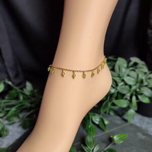 May include: A gold-coloured anklet featuring small, dangling green beads. The delicate chain encircles the ankle, with the green beads evenly spaced. The anklet is displayed on a foot, against a dark backdrop with foliage.