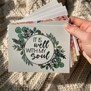 Może przedstawiać: Stos białych kartek z akwarelowymi wzorami kwiatowymi i napisem "IT IS well WITH MY SOUL". Kartki przedstawiają zielone liście i pomarańczowe i czerwone kwiaty. Kartki są trzymane w dłoni na tle teksturowanego, kremowego tła.
