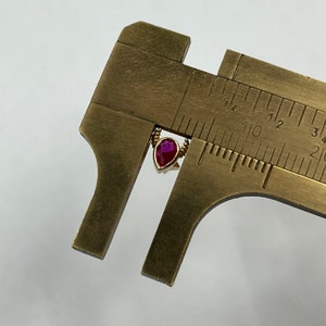 May include: Close-up of a red pear-shaped gemstone pendant measured with a caliper. The pendant is approximately 1/2 inch in length.