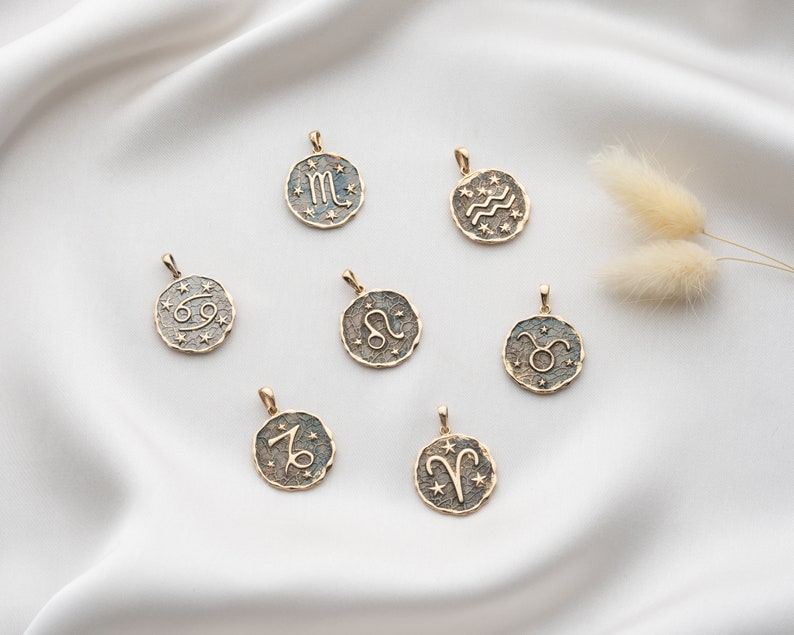 Puede incluir: Un conjunto de colgantes de signo del zod&iacute;aco de color dorado sobre un fondo blanco. Cada colgante presenta un s&iacute;mbolo del zod&iacute;aco diferente, incluidos Aries, Tauro, G&eacute;minis, C&aacute;ncer, Leo, Virgo, Libra, Escorpio, Sagitario, Capricornio, Acuario y Piscis.