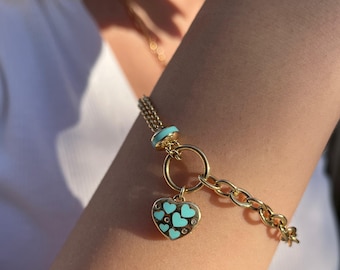 Könnte beinhalten: Ein goldenes Kettenarmband mit einem herzförmigen Anhänger. Der Anhänger ist mit kleineren Herzen verziert und hat eine türkisfarbene Emaille-Oberfläche.