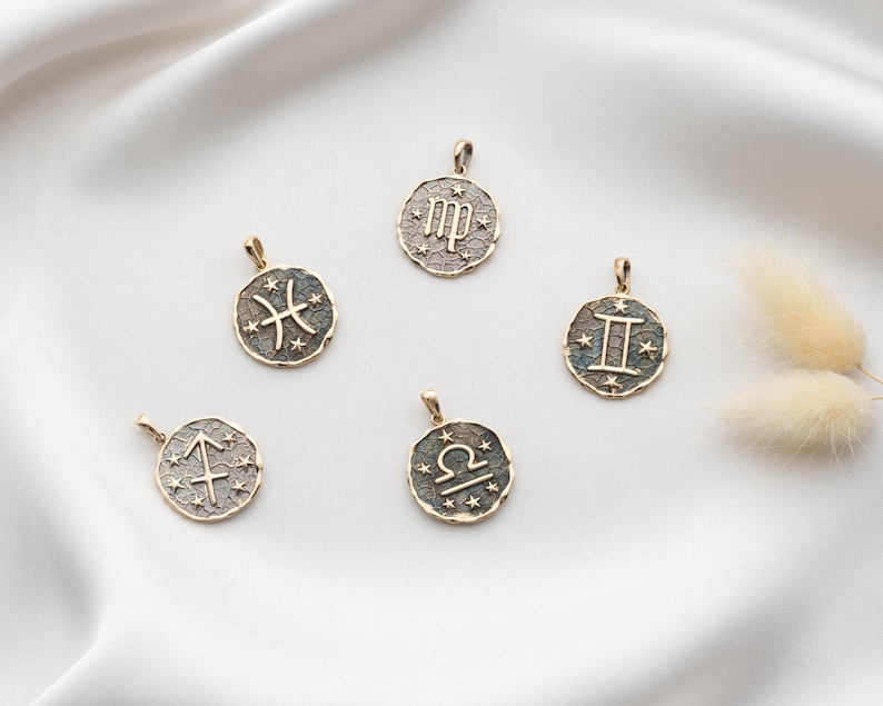 Puede incluir: Cinco colgantes de zodiaco dorados con diferentes signos astrol&oacute;gicos sobre un fondo blanco. Los signos son Piscis, Virgo, G&eacute;minis, Libra y Sagitario.