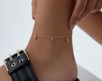 May include: Gold anklet with a delicate chain and three dangling charms. Two charms are gold teardrop shapes, and the third charm is a blue teardrop.