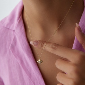 May include: A delicate gold necklace featuring a bee pendant with sparkling accents. The bee pendant is suspended from a fine gold chain. The necklace is displayed against a pink background.