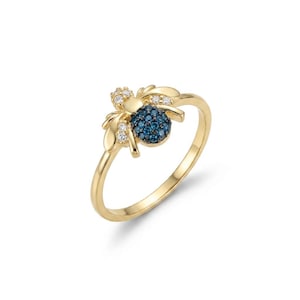 Può includere: Un anello in oro con un elemento centrale a forma di ape. Il corpo dell'ape è incastonato con piccole pietre blu rotonde, mentre le ali sono decorate con minuscole pietre bianche trasparenti. La fascia è realizzata in metallo dorato lucido.