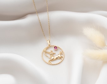 Könnte beinhalten: Goldfarbene Halskette mit einem Anhänger des Sternzeichens Schütze. Der Anhänger zeigt den Schützen mit einem roten Edelstein. Die Halskette hängt an einer feinen Goldkette vor einem weißen Stoffhintergrund.