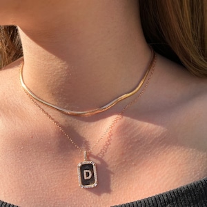 Può includere: Una collana con catena d'oro con un ciondolo con la lettera "D" circondata da piccoli diamanti. Il ciondolo è rettangolare e ha uno sfondo nero.