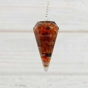 May include: A brown and orange crystal pendulum with a silver chain. The pendulum is hanging from a white wooden surface.