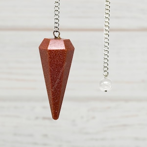 May include: A red gemstone pendulum with a silver chain hanging from a white wooden surface.