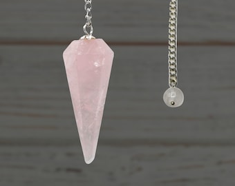 Puede incluir: Un péndulo de cuarzo rosa rosa con una cadena plateada y una pequeña perla blanca en el extremo.