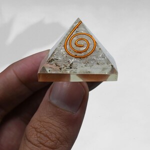 May include: A clear crystal pyramid with a copper wire spiral design. The pyramid is being held by a hand.