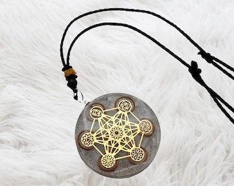 May include: A silver pendant with a gold geometric design and seven brown circles, on a black cord necklace. The pendant is on a white fluffy surface.