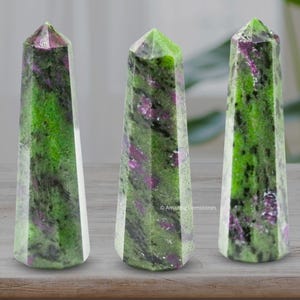 Może przedstawiać: Trzy zielone i fioletowe obeliski z kamieni szlachetnych ze spiczastym wierzchołkiem są wystawione na drewnianej powierzchni. Obeliski mają naturalny, teksturowany wygląd. Tekst nad obeliskami mówi: „Można go umieścić w domu i biurze jako pole magnetyczne”.