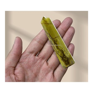 May include: A rectangular, yellow-green gemstone obelisk with brown veining is held in an open hand. The polished stone has a pointed top and a smooth, reflective surface. The background is a neutral beige.