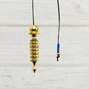 Puede incluir: Un péndulo de color dorado con una punta puntiaguda y una serie de anillos que cuelgan de una cuerda negra sobre un fondo de madera blanca.