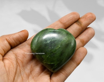 May include: A green heart-shaped stone, also known as a nephrite jade heart, is held in a person's hand. The stone is smooth and polished, with a light green color.