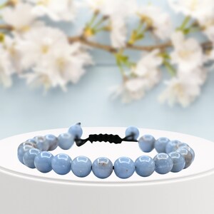 Könnte beinhalten: Ein hellblaues Edelstein-Armband mit einem schwarzen Kordelverschluss. Das Armband besteht aus runden Perlen und wird auf einem weißen Sockel präsentiert.