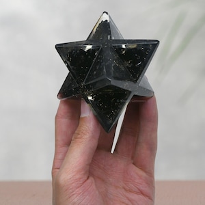 May include: A black obsidian star shaped crystal, also known as a Merkabah, is held in a hand. The crystal is clear with black inclusions.
