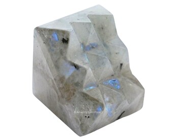 Puede incluir: Un cristal de labradorita blanco facetado e iridiscente con destellos azules y grises. El cristal tiene forma de pirámide con una base plana.