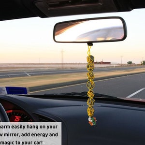 May include: A yellow string with brown crystal beads hangs from the rearview mirror of a car. The car is driving on a highway. The text "Crystal car charm easily hang on your car's rear view mirror, add energy and crystal magic to your car!" is at the bottom of the image.