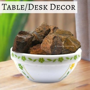 Puede incluir: Un cuenco blanco con un diseño floral verde y amarillo, lleno de piedras ásperas marrones y doradas. El texto "TABLE/DESK DECOR" está en la imagen. Las piedras tienen una apariencia texturizada y varían en tamaño.