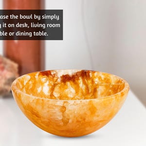 Puede incluir: Un cuenco decorativo con un exterior de color ámbar cálido y una superficie texturizada. El cuenco tiene forma redondeada y está hecho de un material que parece translúcido. El texto de la imagen dice: "Showcase the bowl by simply putting it on desk, living room table or dining table."
