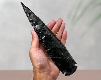Puede incluir: Una punta de flecha de obsidiana negra con una punta puntiaguda y una superficie texturizada. La punta de flecha se sostiene en una mano.