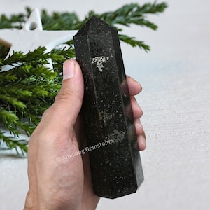 May include: A black obsidian crystal point with a natural, rough texture. The crystal is approximately 15cm long and has a pointed tip. The crystal is held in a hand, with the background blurred.
