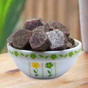 May include: A white bowl with a green and yellow floral design filled with rough, raw, purple ruby gemstones.