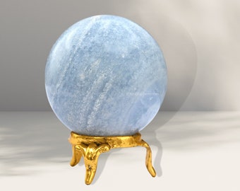 Puede incluir: Una esfera de Celestita azul claro sobre un soporte de metal dorado con tres patas. La esfera es lisa y tiene un aspecto nublado.