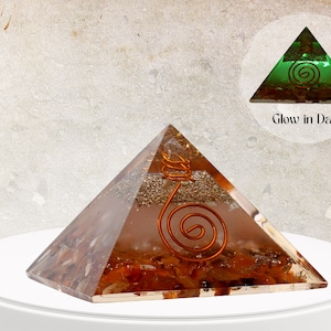 Könnte beinhalten: Eine klare Quarzkristallpyramide mit einer Kupferspirale im Inneren. Die Pyramide ist mit kleinen orangefarbenen und braunen Kristallen gefüllt. Die Pyramide leuchtet im Dunkeln grün. "Glow in Dark" steht unter dem Bild der Pyramide.