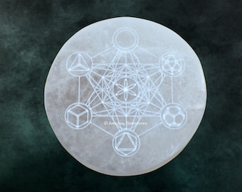 May include: A round, off-white selenite crystal plate with a Metatron's Cube design etched into the surface. The geometric pattern is a series of interconnected shapes and lines. The background is a dark, mottled green.
