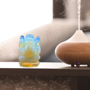 May include: A small, blue and yellow opal Ganesha statue on a wooden table. The statue is sitting in front of a wooden essential oil diffuser.