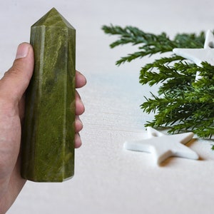 以下が含まれることがあります： 白い背景に、緑の葉と白い星のオーナメントが付いた緑の枝があり、その枝を手に持った人が緑の水晶のポイントを持っている様子です。