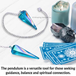Op de afbeelding: Een blauwe, iriserende kristallen slinger met een zilveren ketting die boven een wit houten oppervlak hangt. De slinger wordt omringd door andere waarzegtools, waaronder een kaars, een klein doosje met een kristal erin en een deck tarotkaarten.