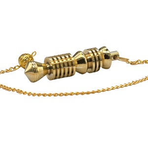 Op de afbeelding: Een gouden slinger met een ketting. De slinger heeft een puntige punt en een cilindrisch lichaam met meerdere ringen. De ketting is bevestigd aan een kleine bal aan de bovenkant.