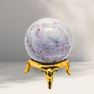 Puede incluir: Una esfera blanca y morada con un patrón de remolino se asienta sobre un soporte de metal dorado con tres patas. "Amazing Gemstones" está impreso en el soporte.