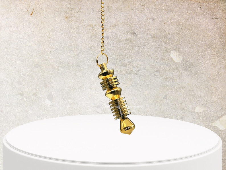 Op de afbeelding: Een gouden metalen slinger met een ketting die aan een wit oppervlak hangt.