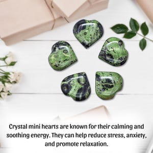 Puede incluir: Cinco cristales en forma de coraz&oacute;n con un patr&oacute;n verde y negro est&aacute;n dispuestos sobre una superficie de madera blanca. Los cristales est&aacute;n rodeados de hojas verdes y una caja de regalo. El texto de la imagen dice: "Los mini corazones de cristal son conocidos por su energ&iacute;a calmante."