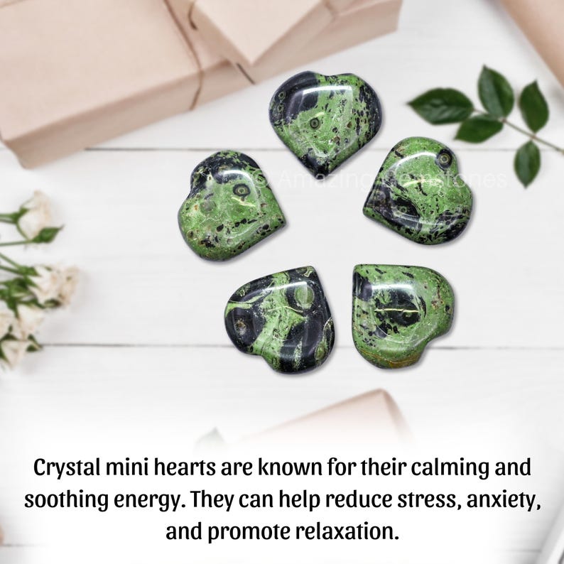 Puede incluir: Cinco cristales en forma de coraz&oacute;n con un patr&oacute;n verde y negro est&aacute;n dispuestos sobre una superficie de madera blanca. Los cristales est&aacute;n rodeados de hojas verdes y una caja de regalo. El texto de la imagen dice: "Los mini corazones de cristal son conocidos por su energ&iacute;a calmante."