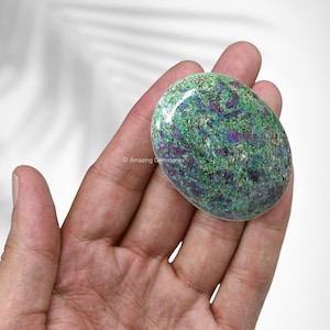 May include: A green and purple ruby zoisite gemstone, also known as ruby in zoisite, in a tumbled oval shape. The stone has a smooth, polished surface and is speckled with red and purple inclusions.