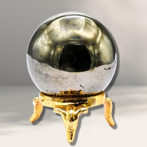 Op de afbeelding: Een grote, ronde, zilveren pyrietbol op een gouden metalen standaard met drie poten. De bol is hoogglanzend gepolijst en reflecteert licht.